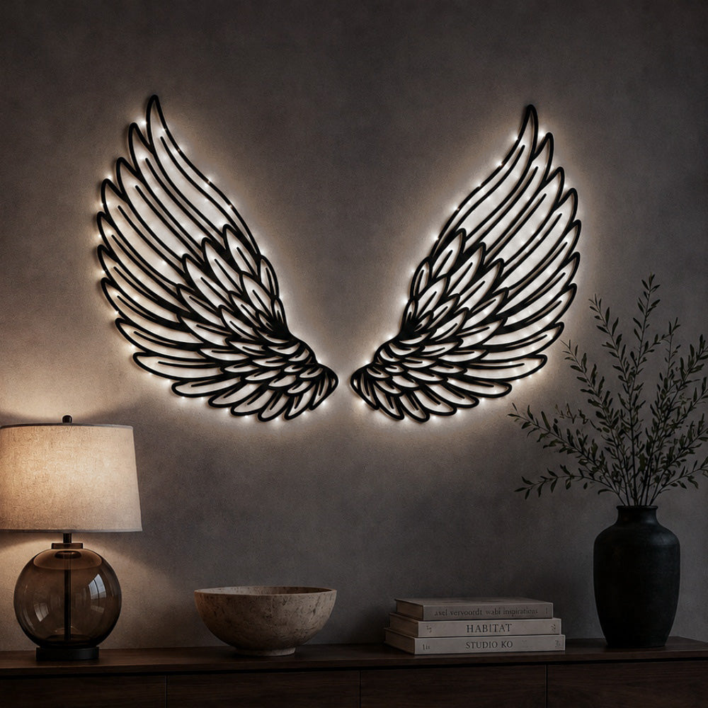 A pair of black metal wing wall art pieces with warm white LED backlighting, mounted on a textured beige wall above a wooden console table with a lamp, bowl, books, and a vase